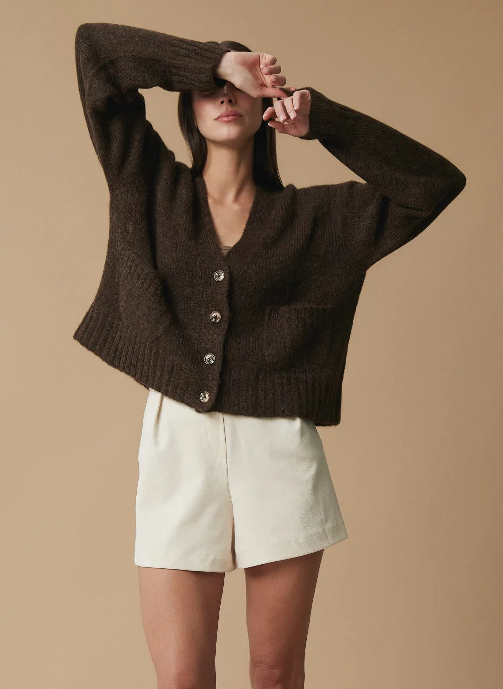 A woman modeling a dark brown chunky knit alpaca cardigan with silver buttons, paired with cream shorts, against a solid beige background.