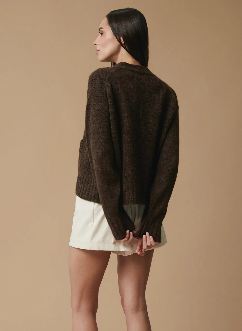 A woman shown from the back wearing a dark brown knit sweater and off-white shorts against a solid beige studio background.