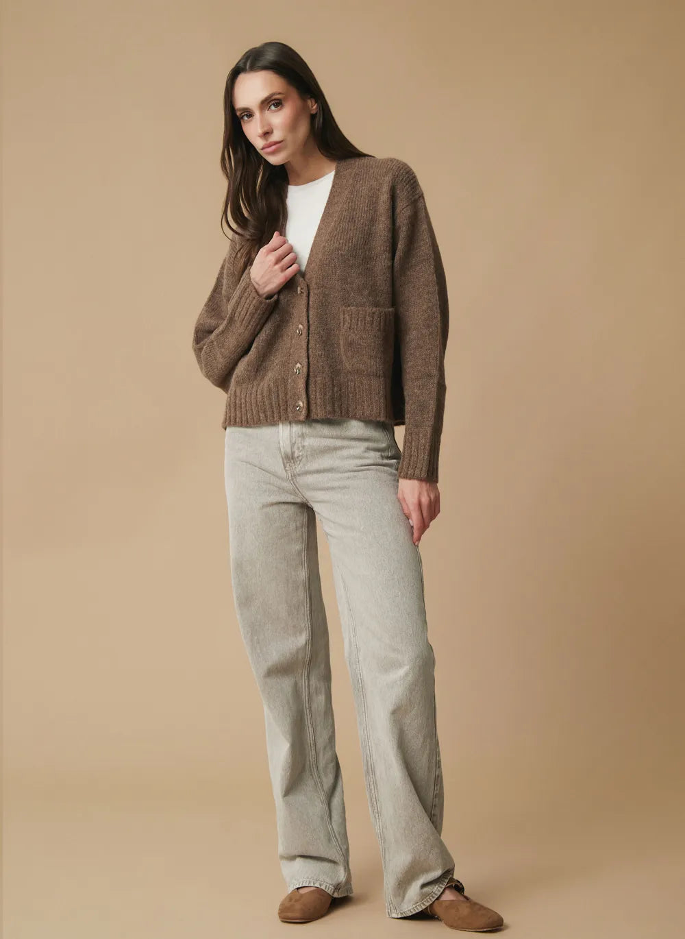 A woman modeling a alpaca brown knit cardigan over a white top, paired with light gray wide-leg jeans and brown shoes, against a solid beige studio background.