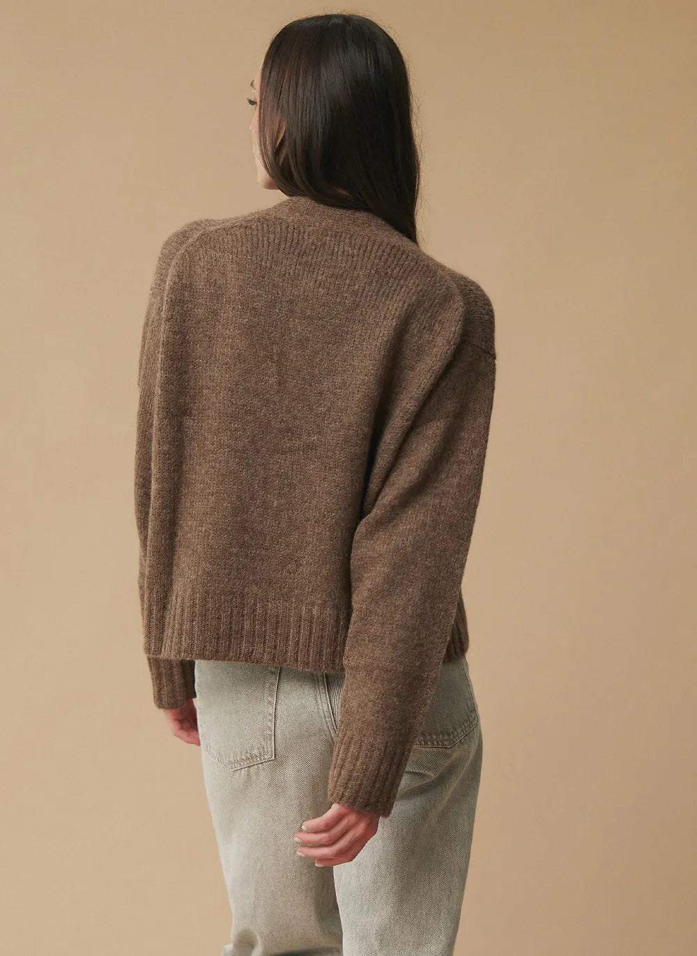 A woman shown from the back wearing a brown ribbed knit cropped sweater and light gray jeans, standing against a plain beige studio background.