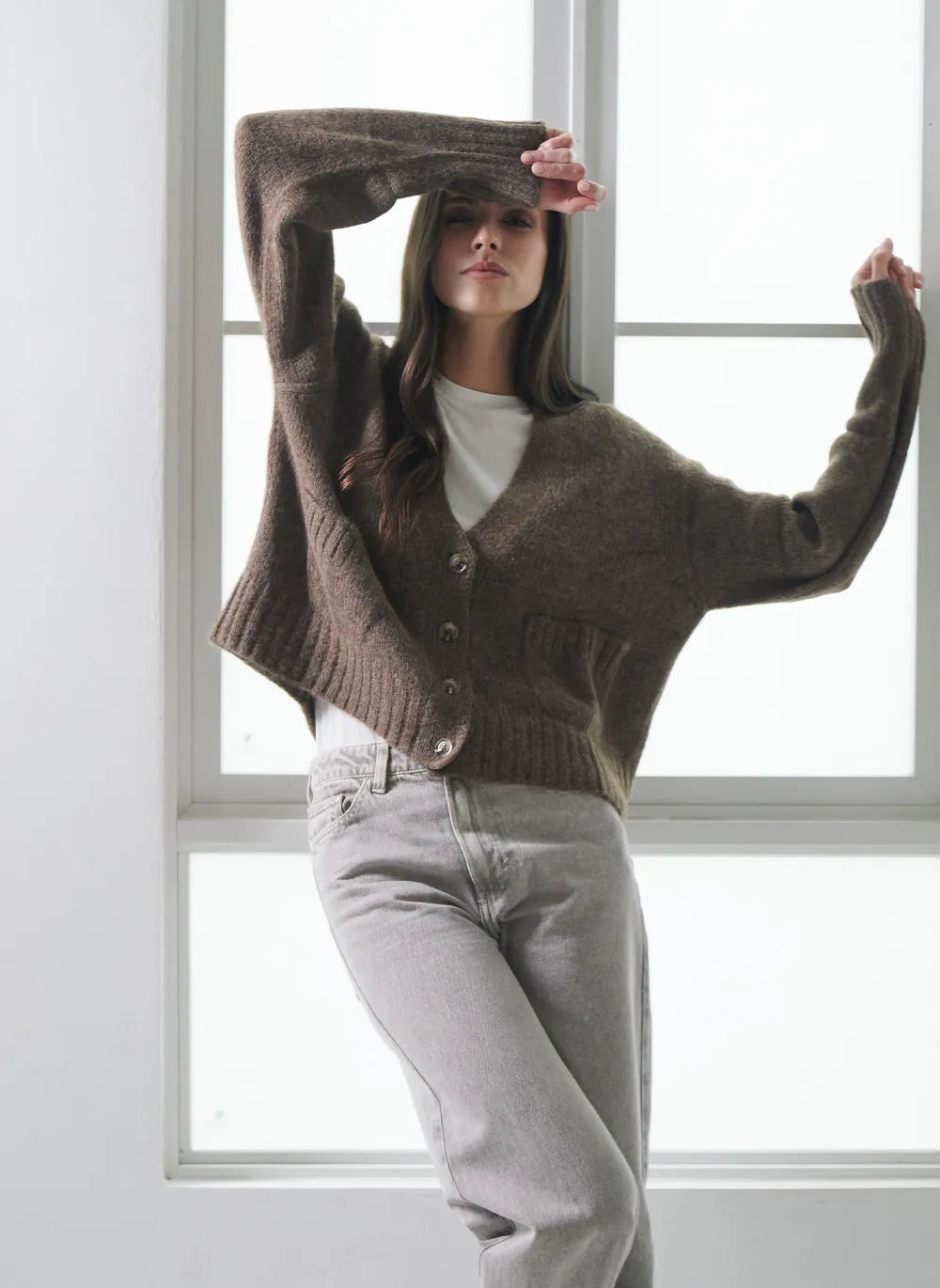 A woman modeling a cropped, oversized brown knit alpaca cardigan sweater with front buttons, worn over a white top and light gray jeans, posing in front of a bright window.