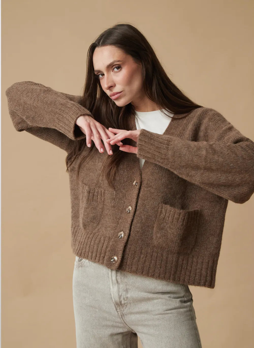 A woman modeling a brown chunky knit cardigan with front buttons and patch pockets, worn over a white top and light gray jeans, against a solid beige studio background.