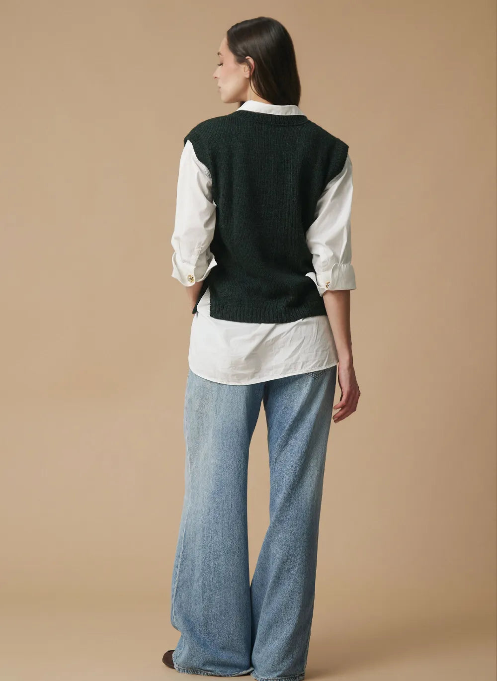 A model is shown from the back wearing a alpaca dark green knit vest layered over a white button-down shirt and paired with wide-leg light blue jeans, set against a solid beige studio background.