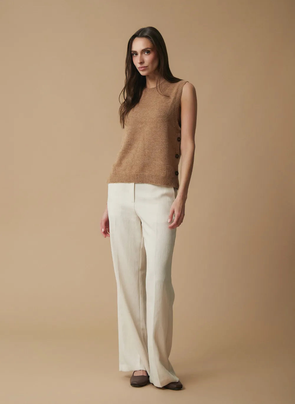 A woman modeling a sleeveless tan knit sweater vest and off-white wide-leg trousers against a solid beige studio background.