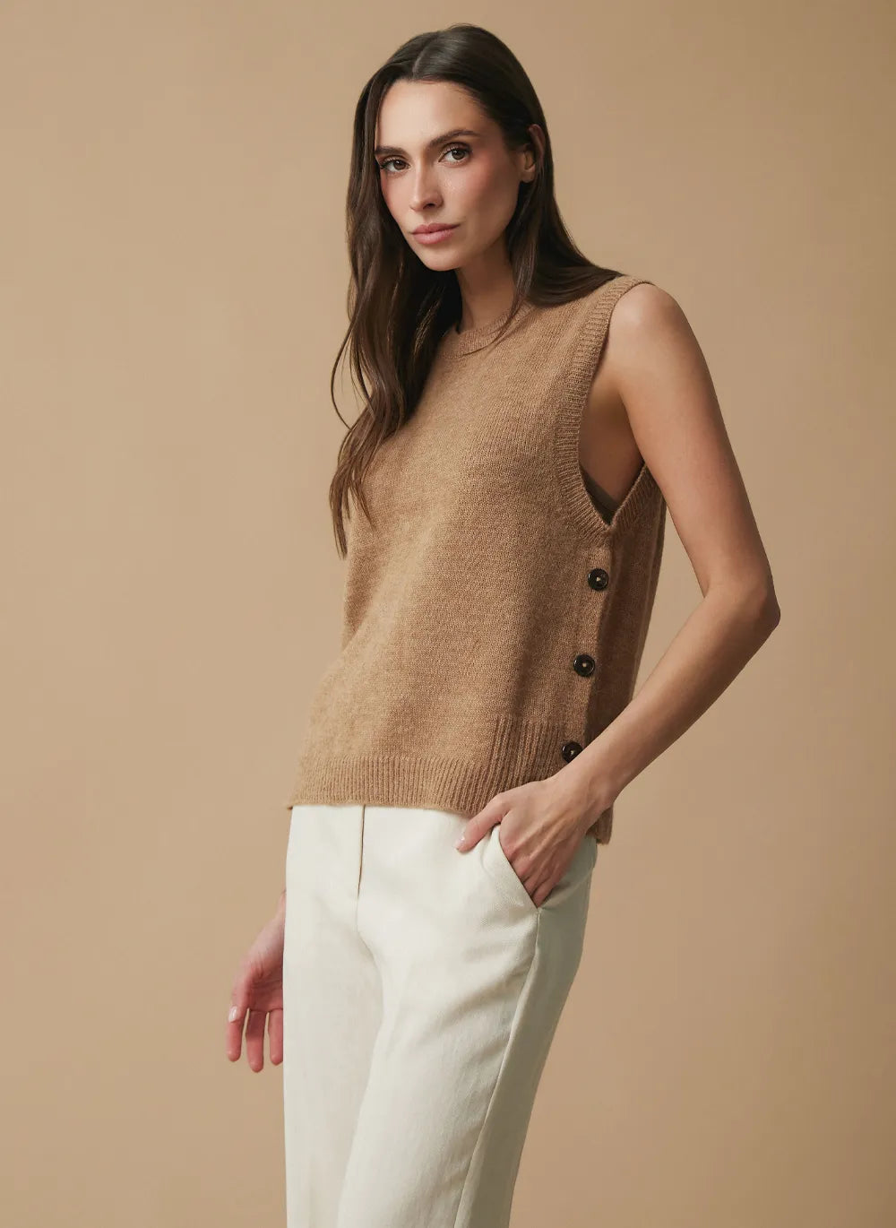 A woman modeling a sleeveless knit vest in a warm camel brown color, styled with white trousers against a solid beige studio background.