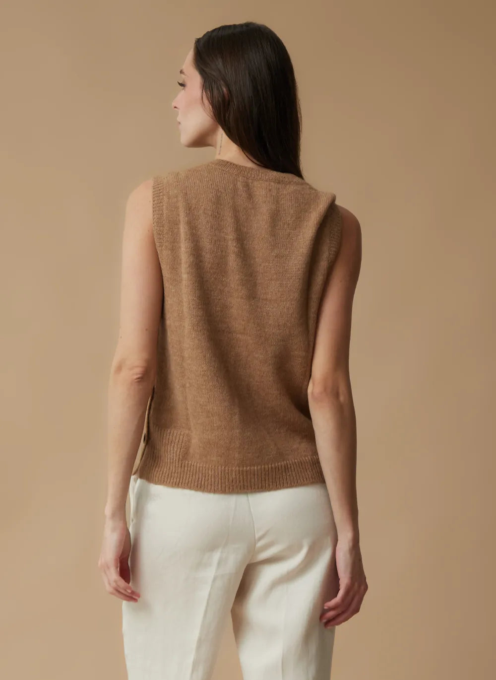 A woman shown from the back wearing a sleeveless camel brown knit sweater vest and off-white trousers, set against a solid warm beige studio background.