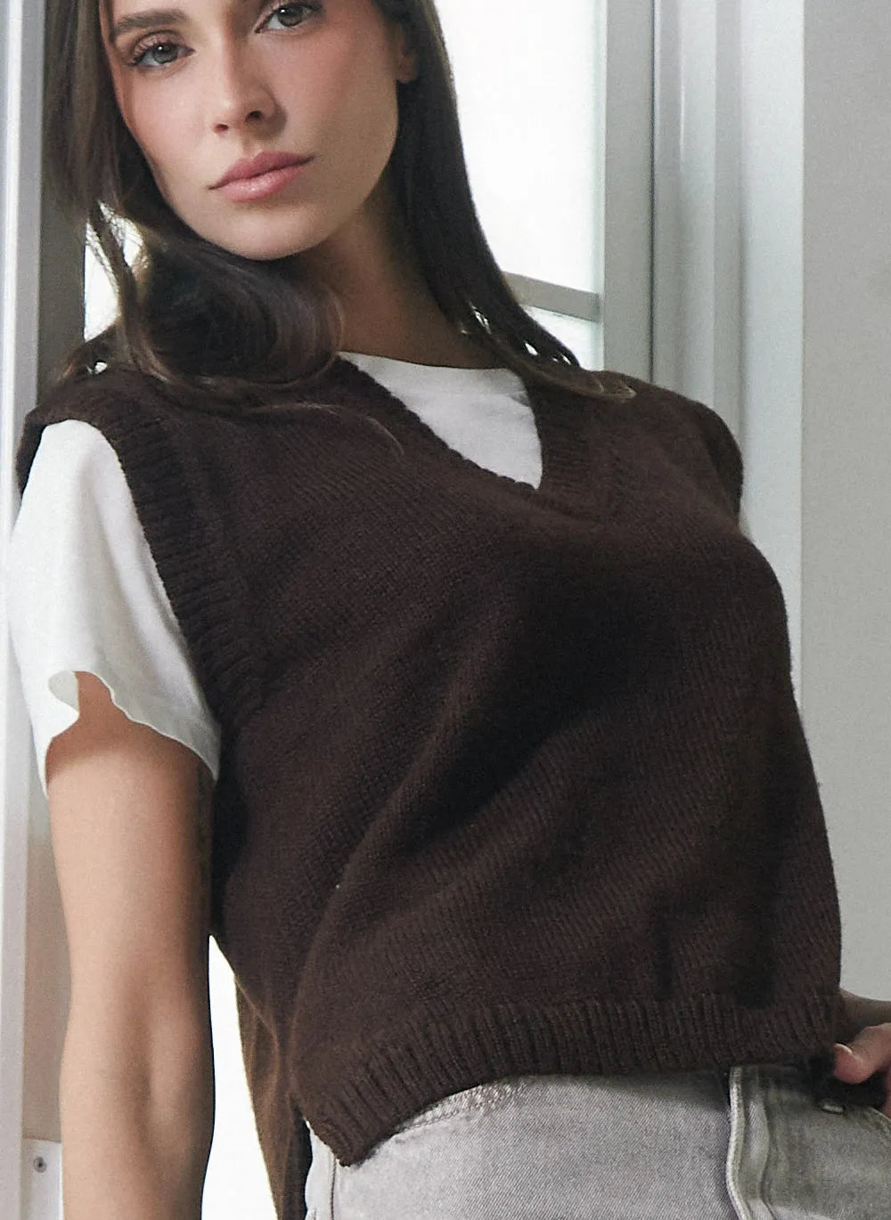 A woman wearing a dark brown ribbed knit sweater vest over a white short-sleeve t-shirt and light gray jeans, standing in a brightly lit indoor setting.