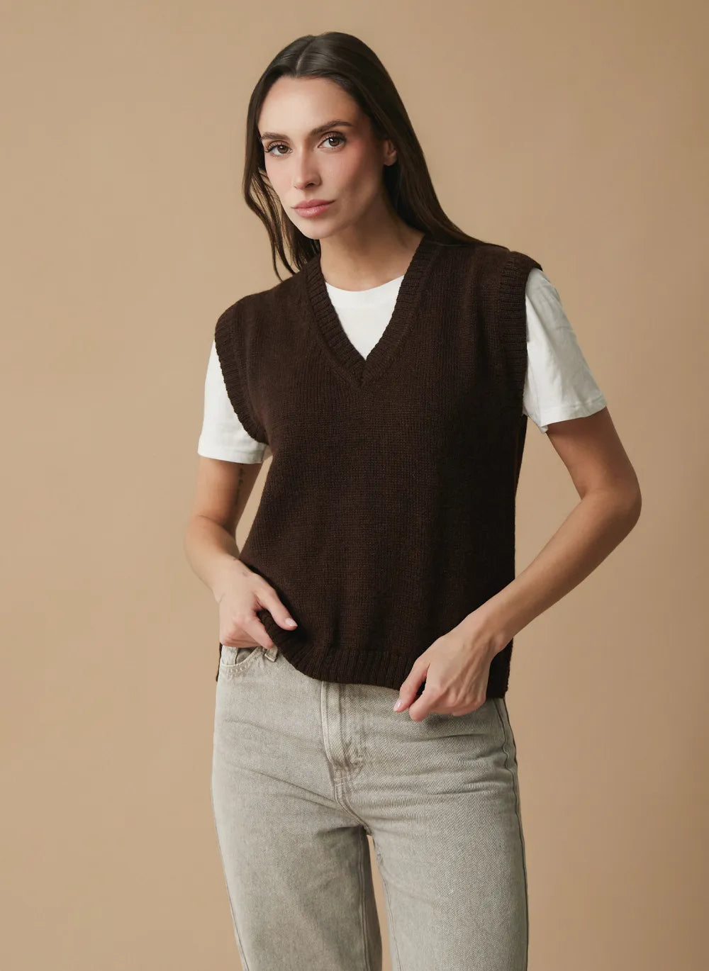 A woman modeling a dark brown knit V-neck sweater vest over a white short-sleeve t-shirt and light gray jeans, set against a solid beige studio background.