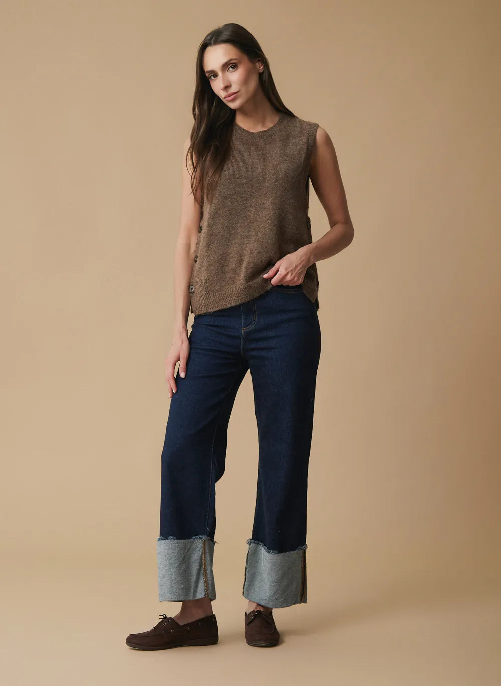 A woman modeling a sleeveless brown knit top, dark blue wide-leg jeans with rolled light blue cuffs, and brown shoes against a solid beige studio background.