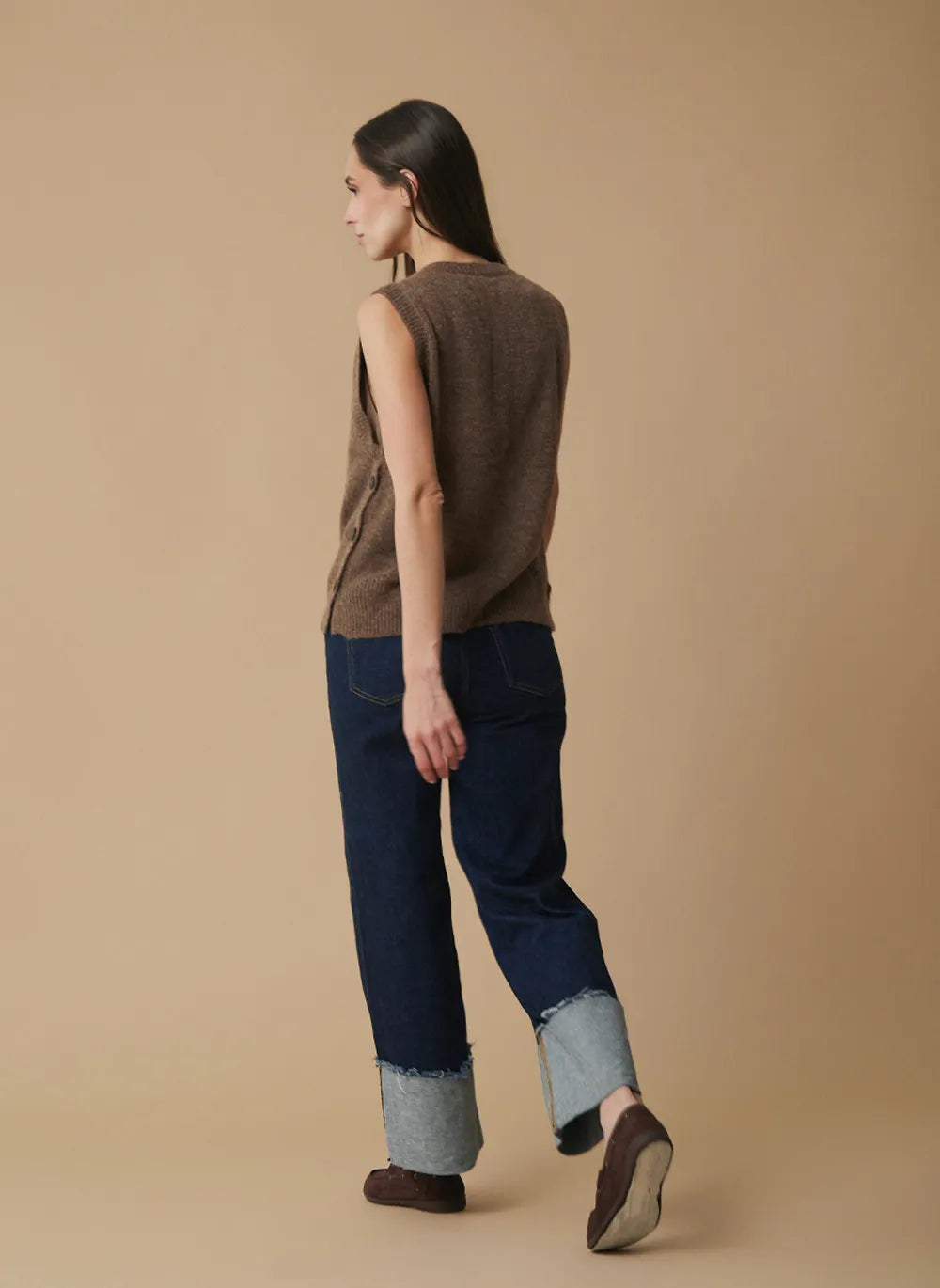 A woman is shown from the back wearing a brown sleeveless knit vest, dark blue cropped jeans with light blue frayed cuffs, and brown slip-on shoes, set against a solid beige studio background.