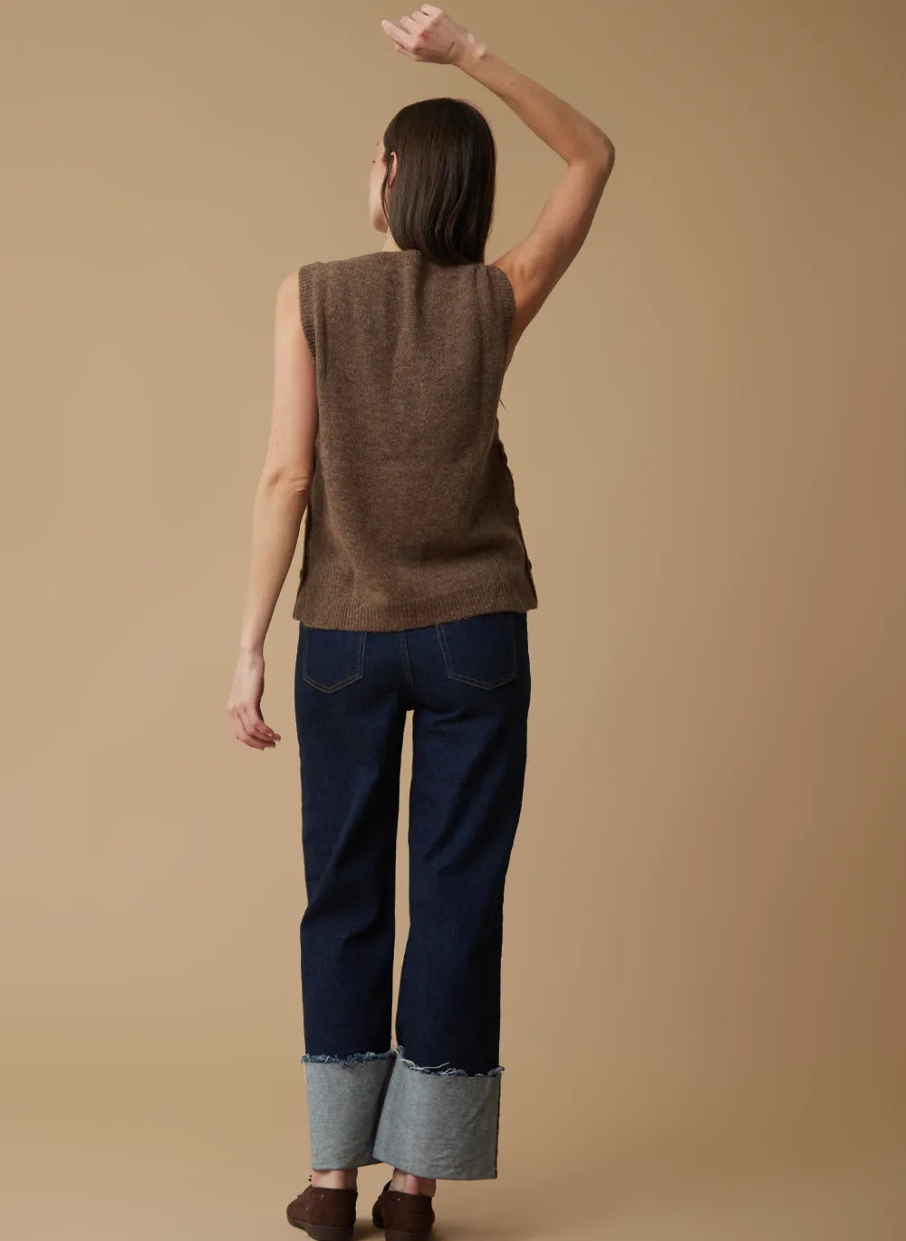 A woman shown from the back wearing a brown sleeveless knit vest, dark blue jeans with light gray rolled cuffs, and brown shoes, against a solid beige studio background.