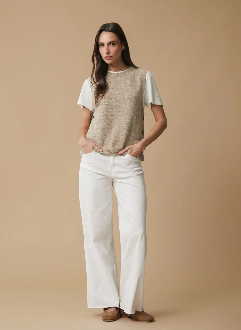 A woman stands against a plain beige studio background wearing a cream short-sleeve t-shirt layered under a sleeveless beige knit vest, paired with white wide-leg pants and brown shoes.