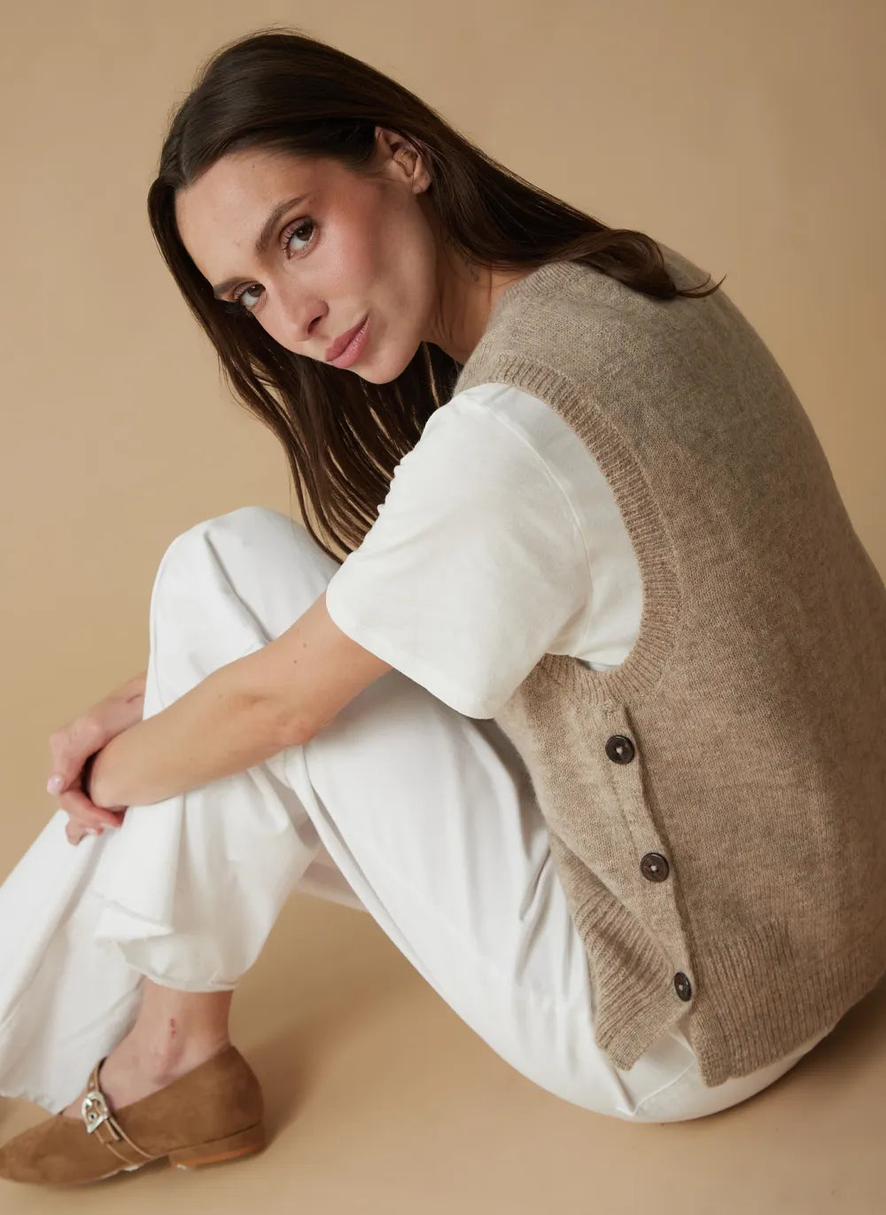 A woman sits cross-legged on a warm beige studio background, wearing a taupe knit button-front vest over a white short-sleeve top and white pants, with tan flats.