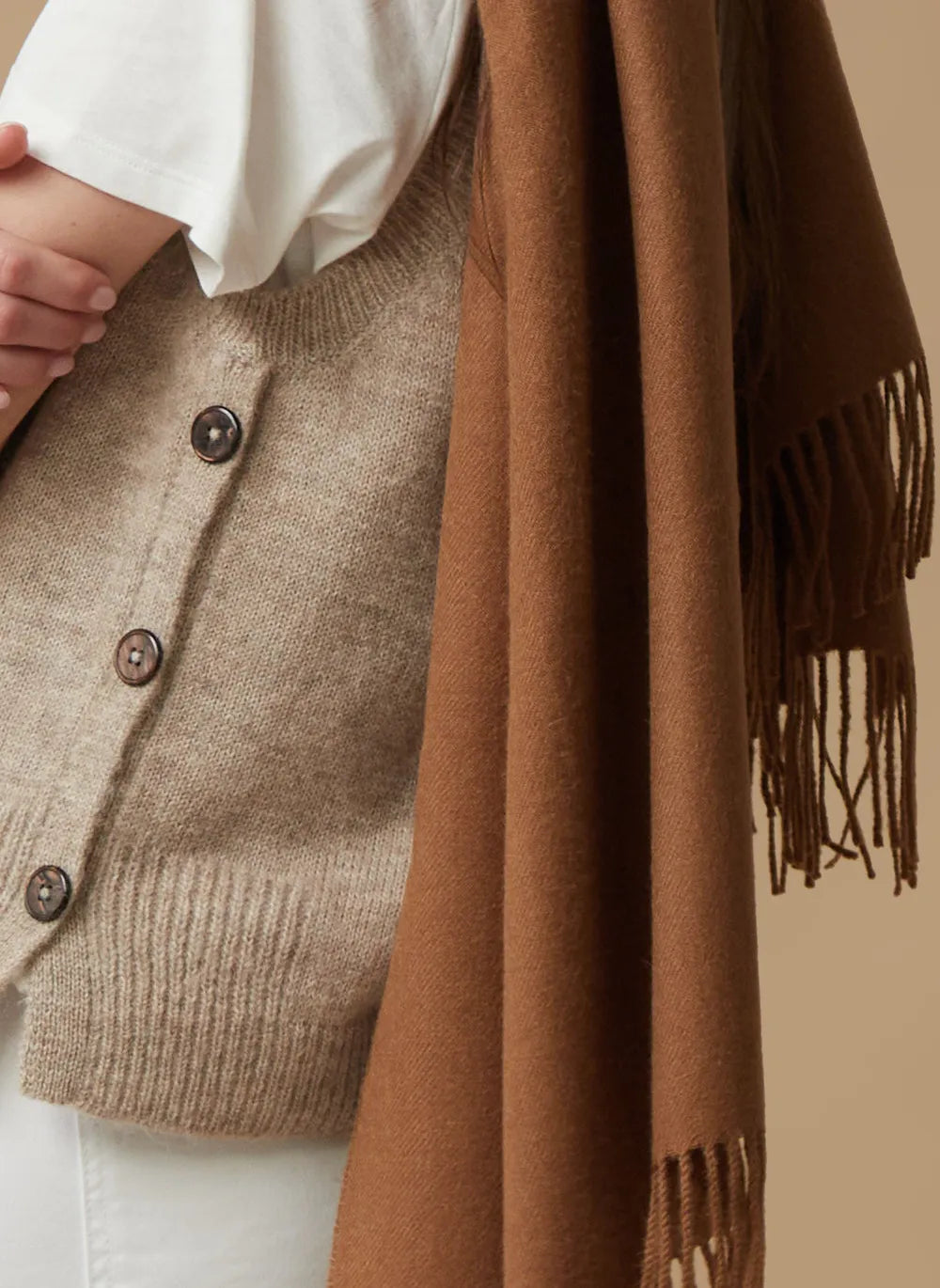 A close-up of a person wearing a beige knit button-front cardigan over a white top, with a rich brown fringed scarf draped over the shoulder against a warm beige background.