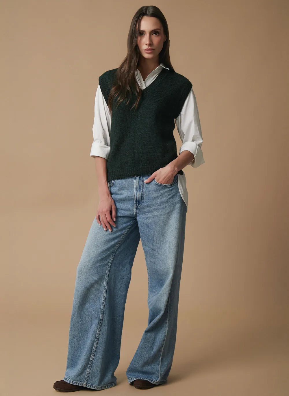 A woman modeling a dark green knit alpaca sweater vest over a white collared shirt and light blue wide-leg jeans, standing against a solid beige studio background.