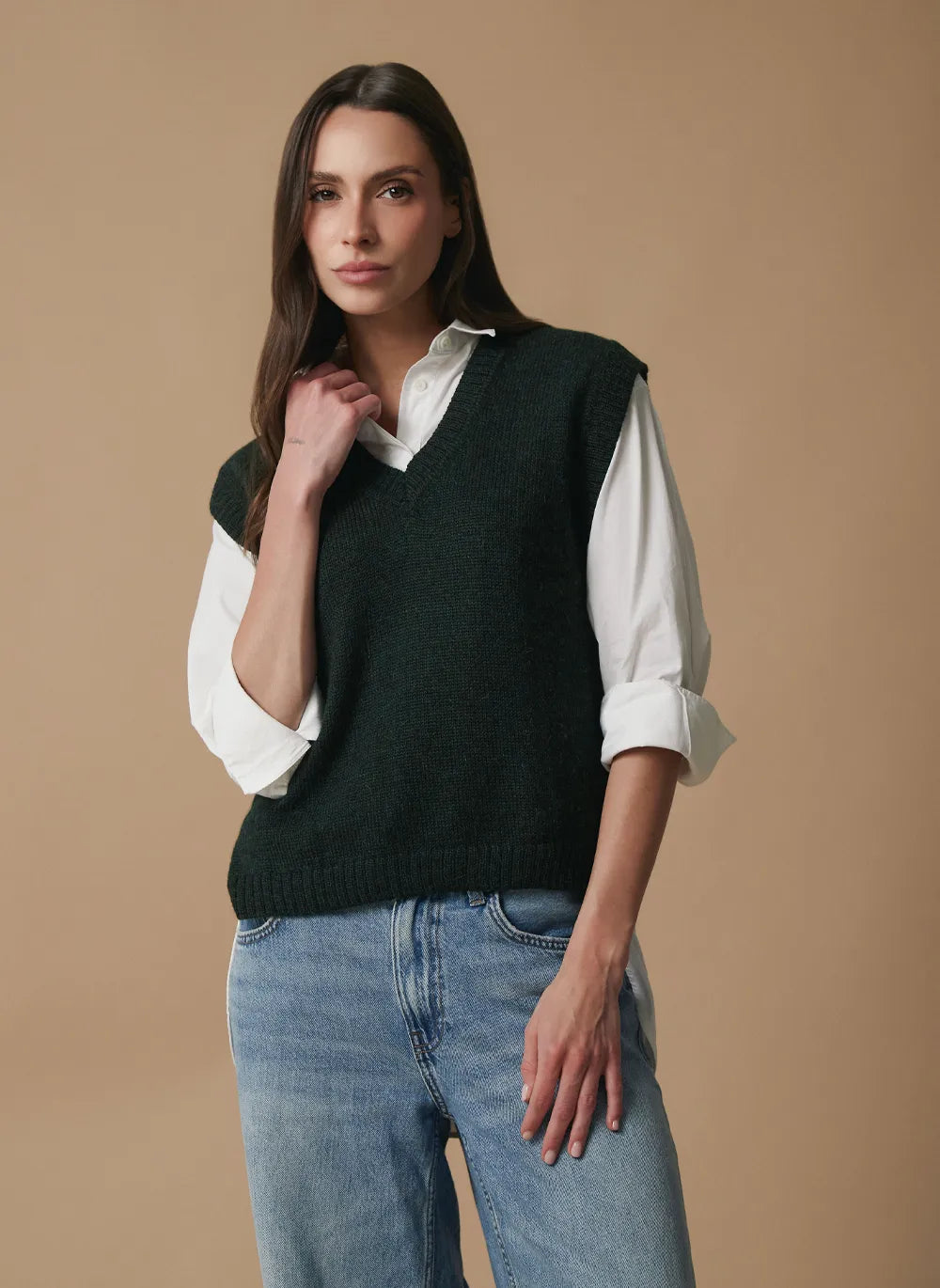 A woman modeling a dark forest green knit sweater vest over a white button-down shirt and blue jeans, set against a solid beige studio background.