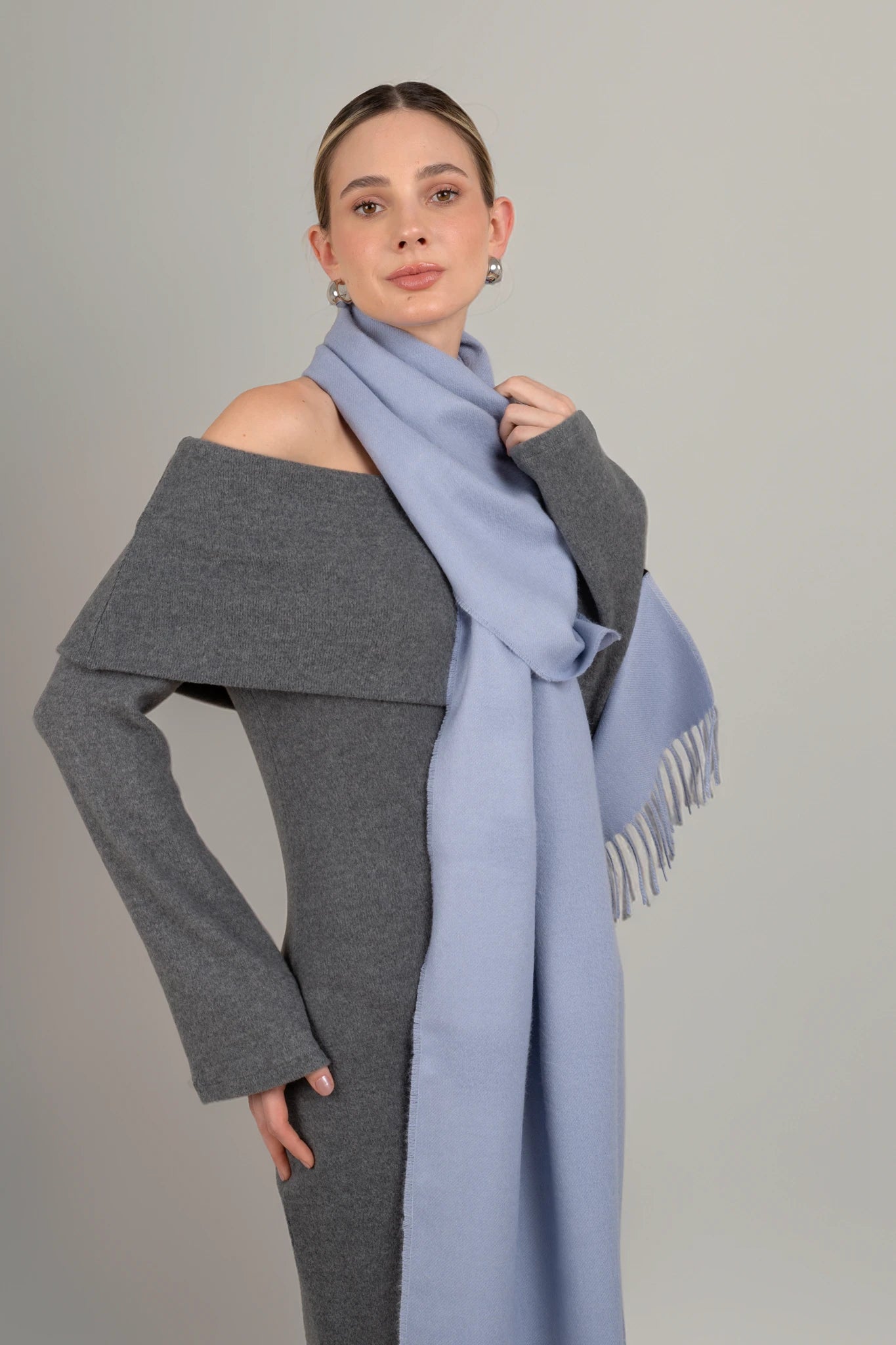 Woman wearing a gray off-shoulder top and a long light blue scarf against a plain background