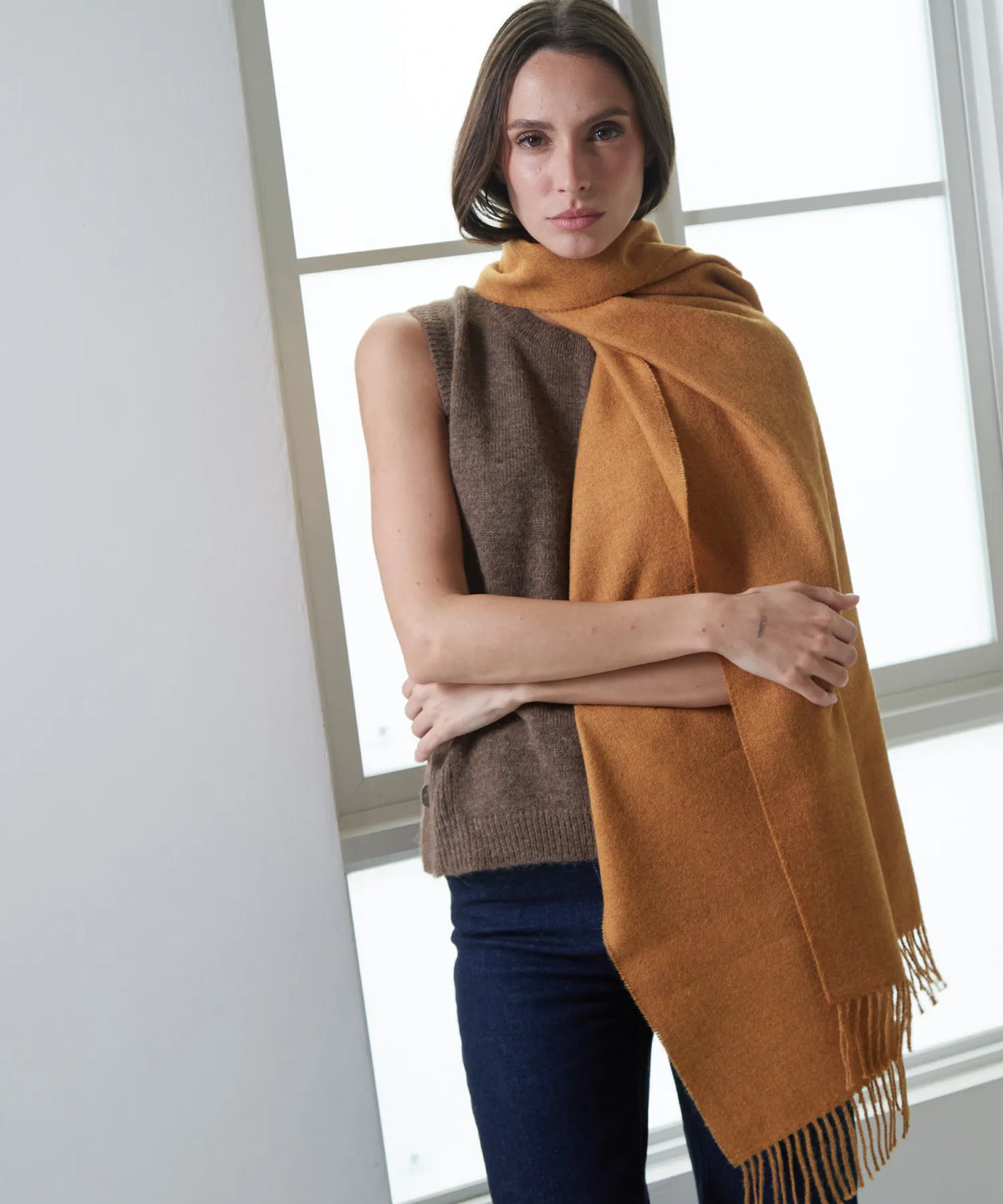 A woman wearing a brown knit sleeveless top and dark blue jeans, wrapped in a large mustard yellow and brown knit scarf with fringe, standing in front of a bright window.