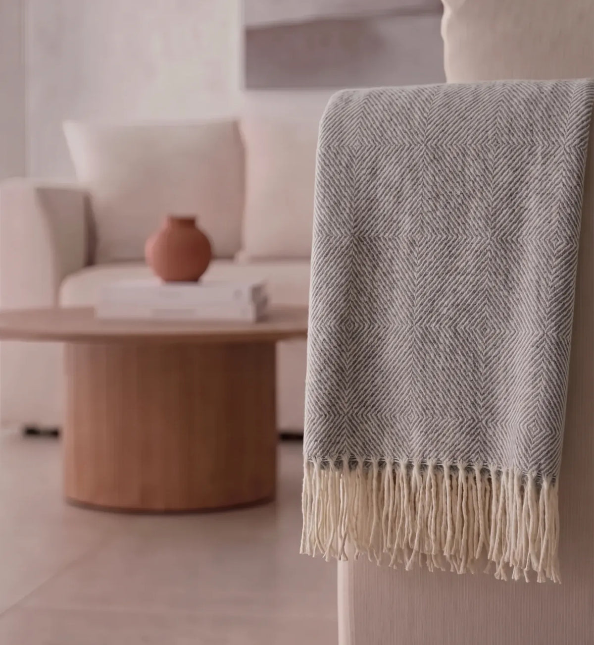 A gray and white herringbone patterned throw blanket with cream fringe, draped over a chair in a softly lit living room.
