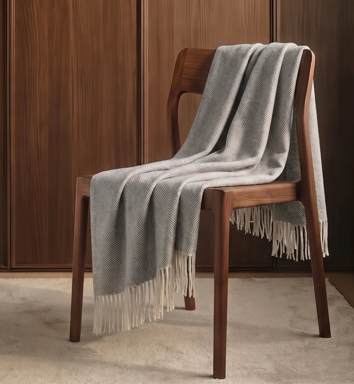 A light gray and white textured knit alpaca throw blanket with cream fringe, draped over a dark brown wooden chair in a warm, modern room setting.