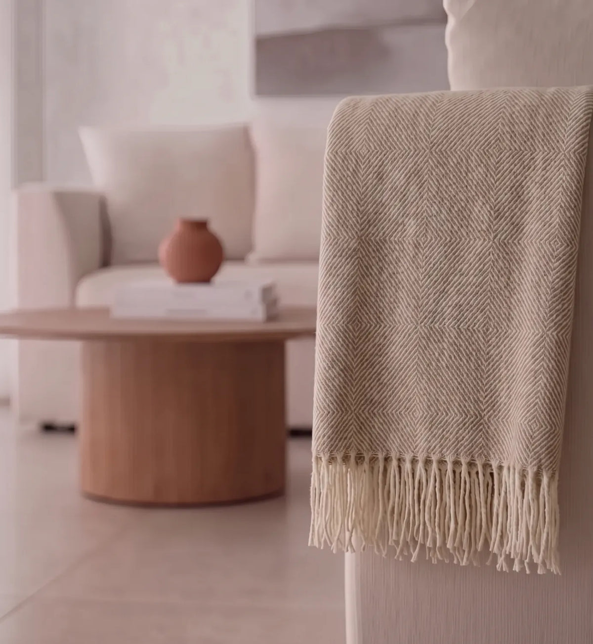 A beige woven throw blanket with a herringbone pattern and fringed edge draped over the back of a sofa in a softly lit living room.