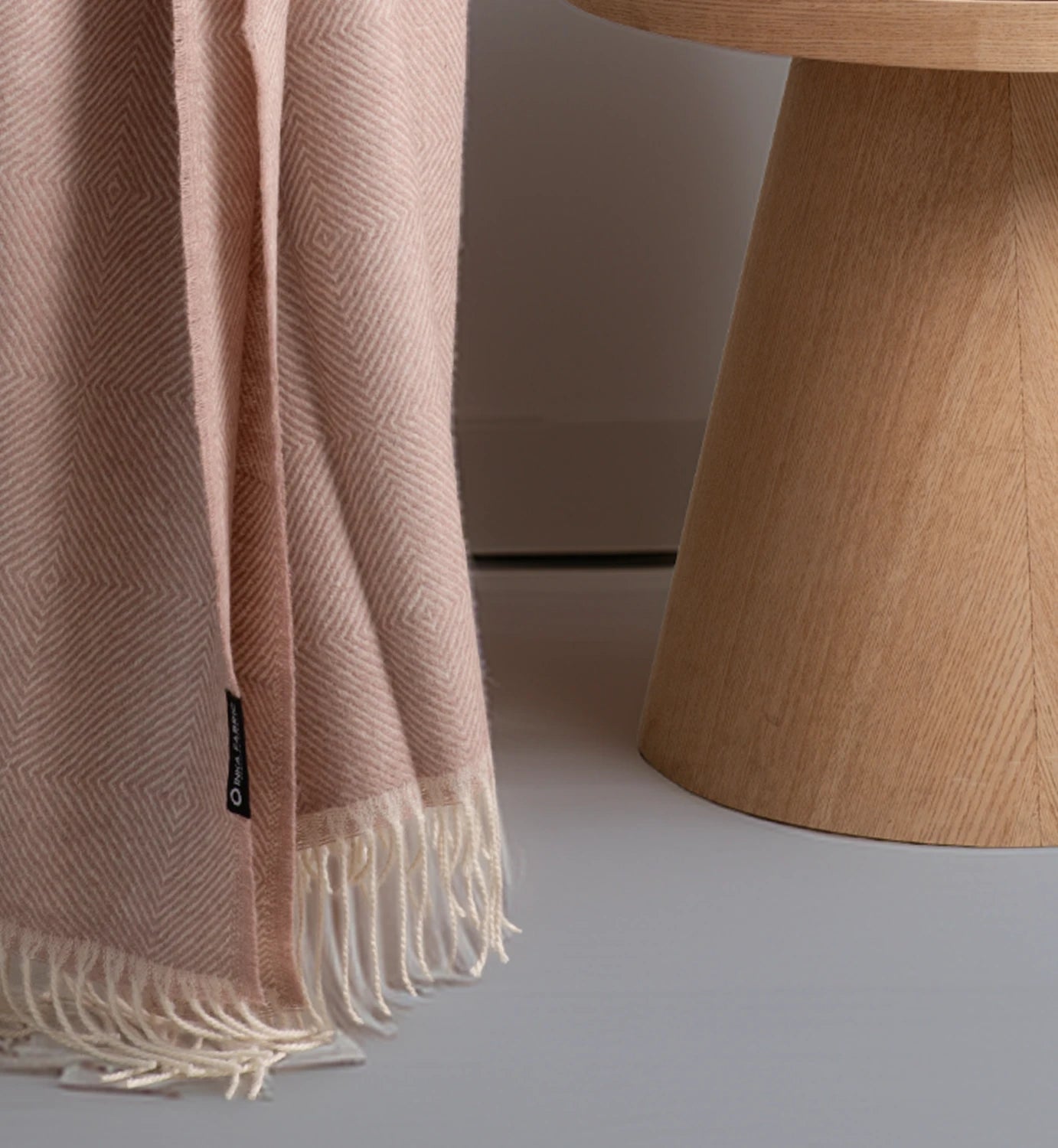 Pink woven blanket with white fringe draped over a wooden stool on a neutral background