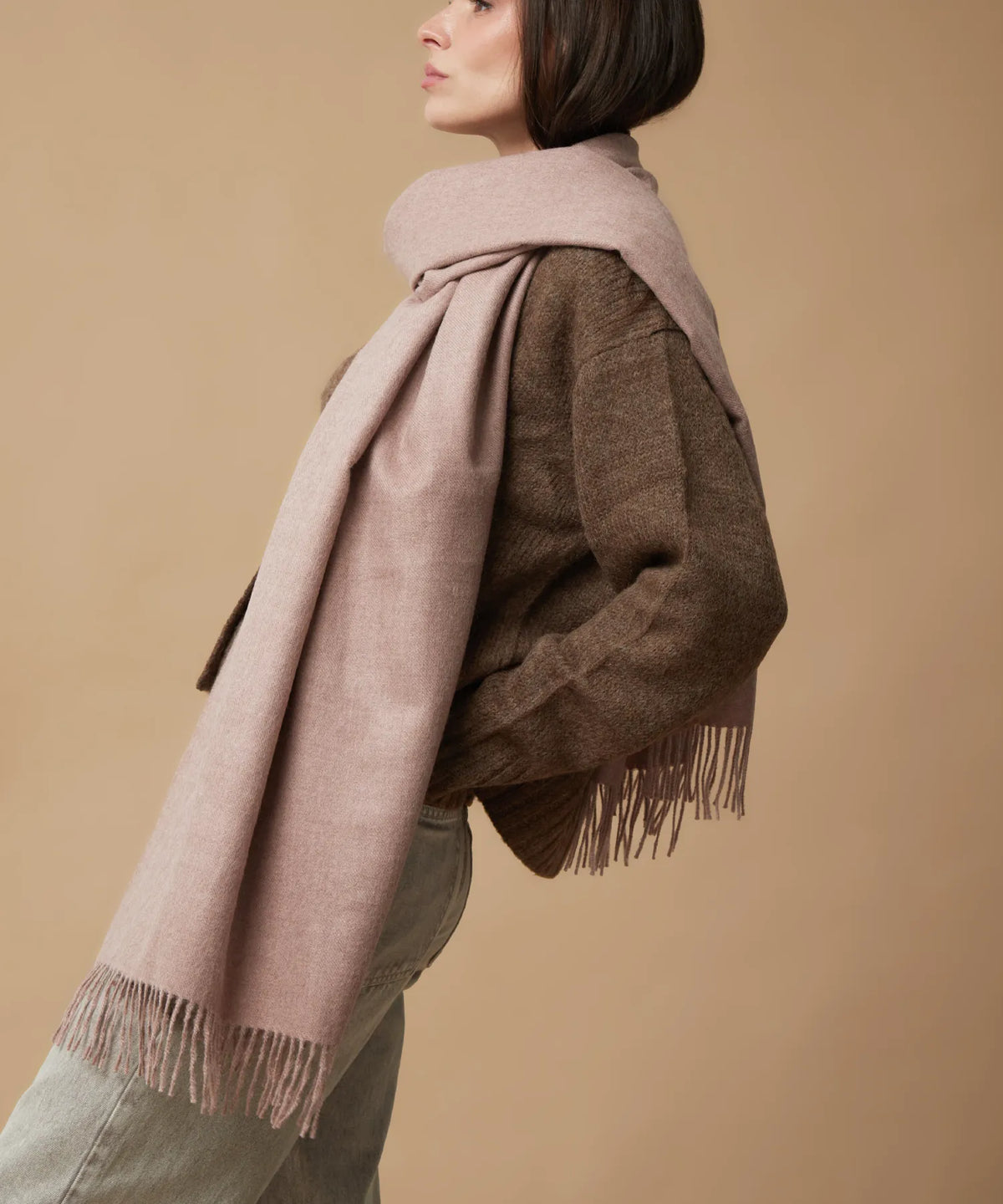 A woman shown from the side wearing a dusty pink knit scarf draped over a brown textured sweater and light olive green jeans, set against a solid beige studio background.