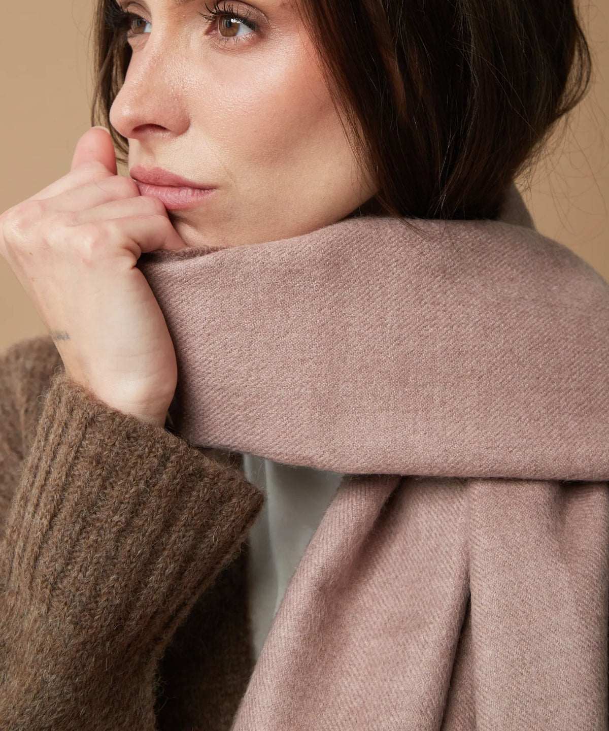 A close-up lifestyle shot of a woman wearing a thick, soft dusty pink scarf wrapped around her neck, paired with a brown ribbed knit sweater against a warm beige background.