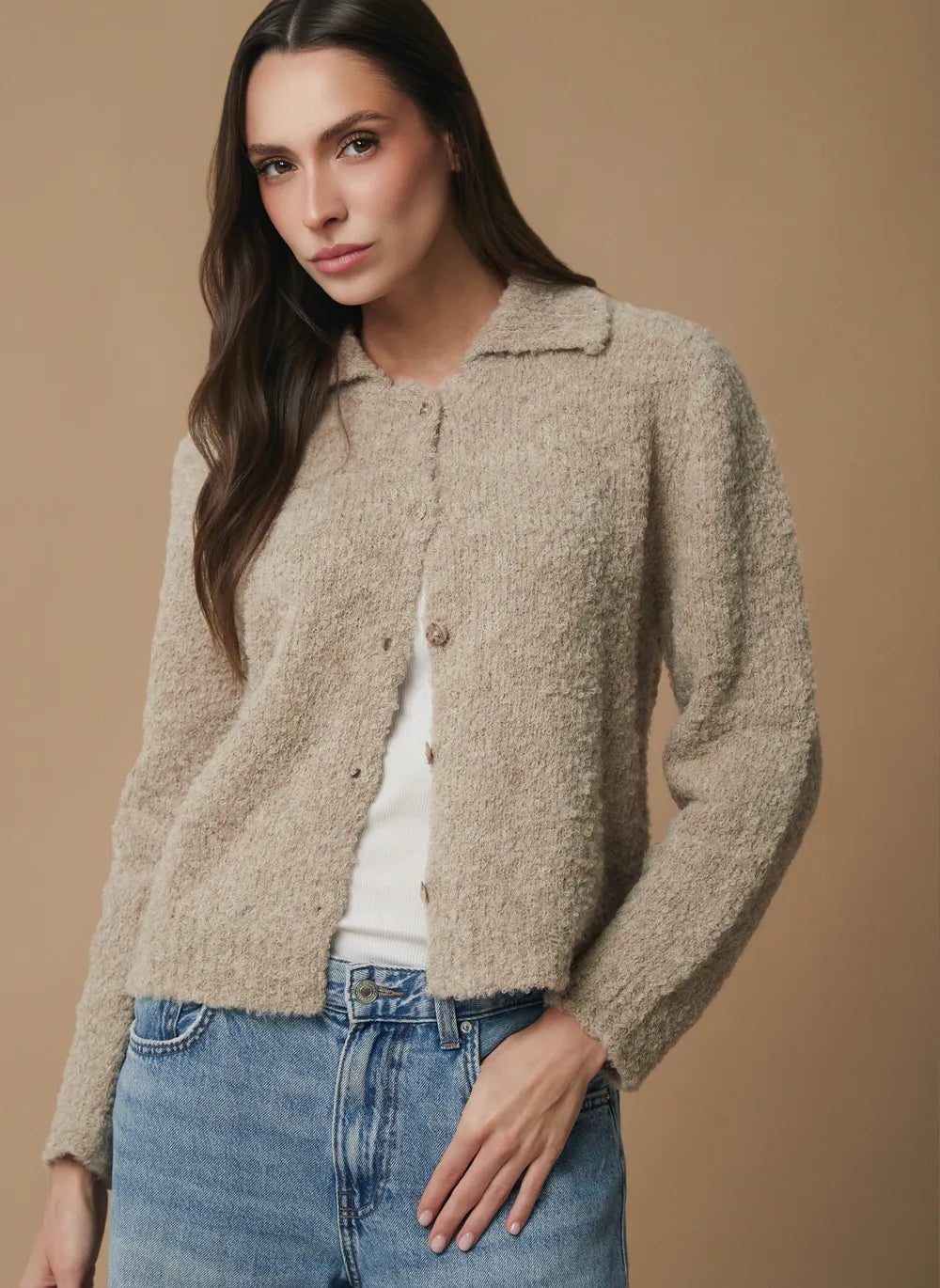 A woman modeling a cropped, textured knit alpaca cardigan in a warm taupe-beige color, worn over a white top and blue jeans against a solid tan studio background.
