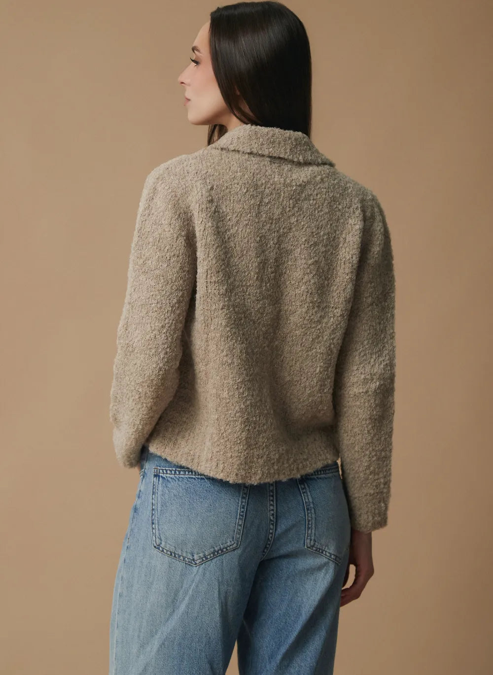 A woman shown from the back wearing a textured, fuzzy taupe-beige cropped cardigan and light blue jeans, set against a solid warm beige studio background.