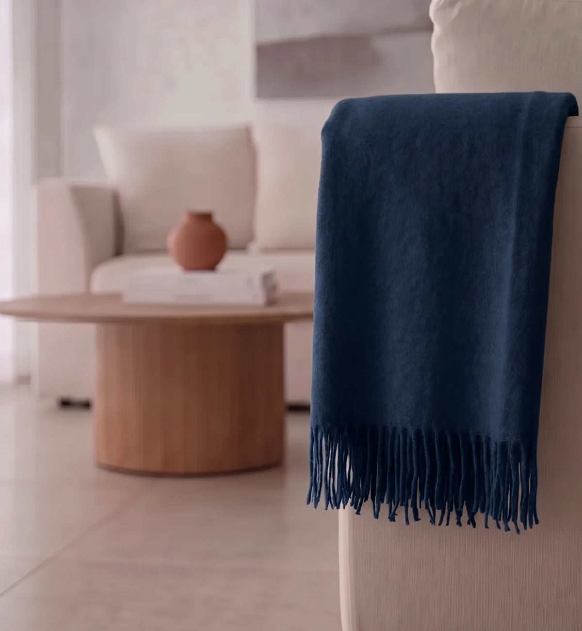 A navy blue throw blanket with fringe trim draped over the arm of a light beige armchair in a softly lit, modern living room.