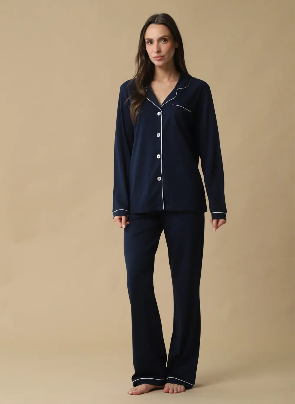A woman modeling a navy blue pajama set with white piping details, consisting of a long-sleeve button-up top and matching pants, against a solid beige background.