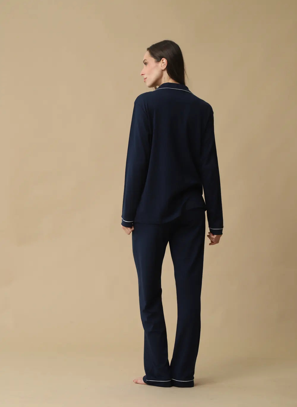 A woman stands with her back to the camera wearing a dark navy blue pajama set with thin white piping trim on the collar, cuffs, and hem, against a solid beige studio background.