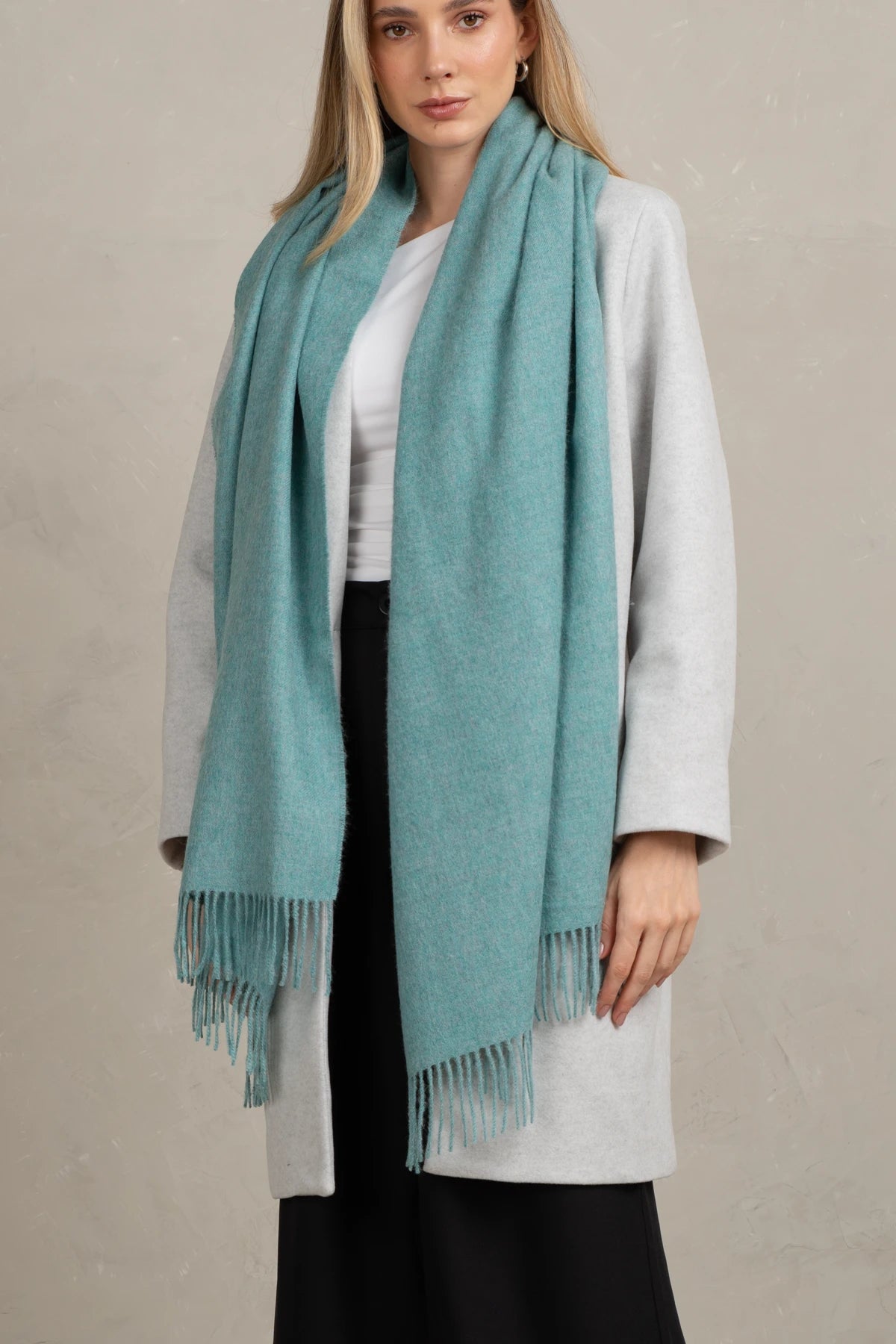 Woman wearing a alpaca wool light blue scarf with fringe, white shirt, and black pants against a beige background