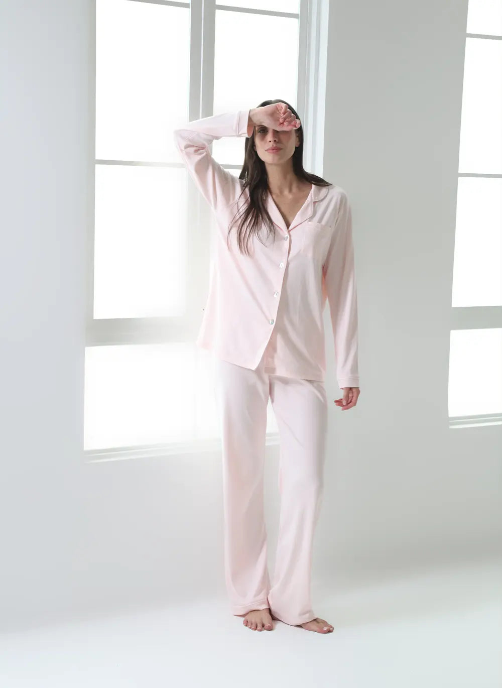 A woman wearing a light pink two-piece pajama set with a long-sleeve button-up top and matching wide-leg pants, standing barefoot in a bright white room with large windows.