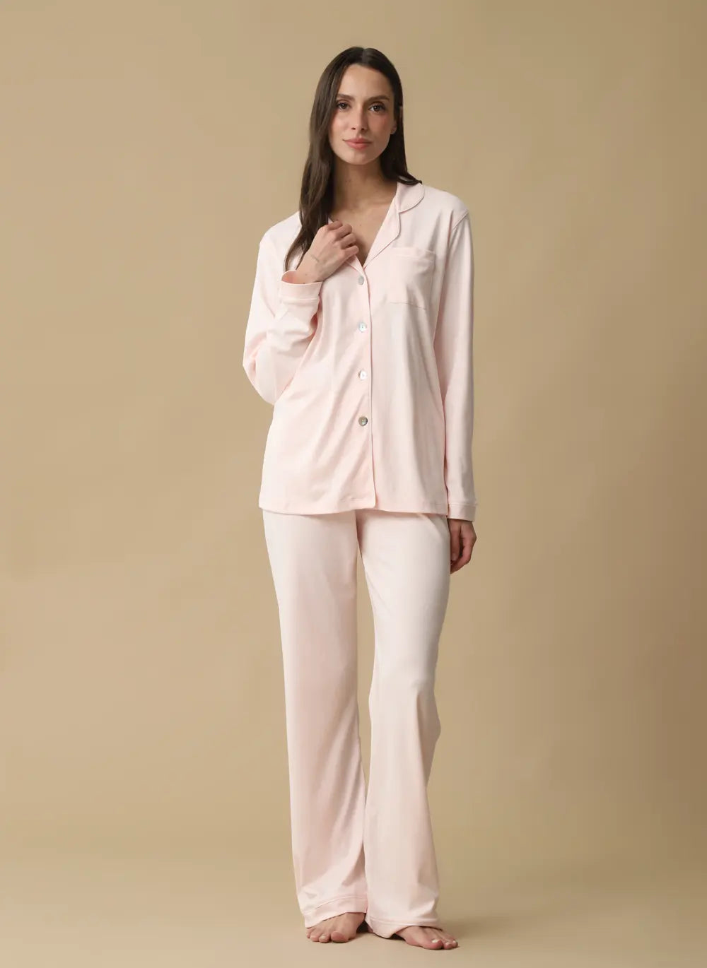 A woman modeling a light pink pajama set consisting of a long-sleeve button-front top and matching wide-leg pants, photographed against a solid beige studio background.