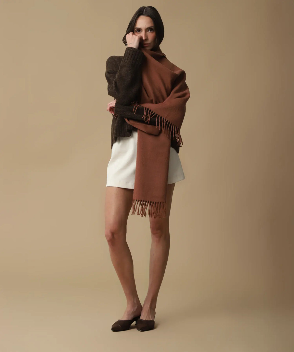 A woman models a rust-brown fringed wrap shawl over a dark brown sweater and white shorts against a solid beige studio background.