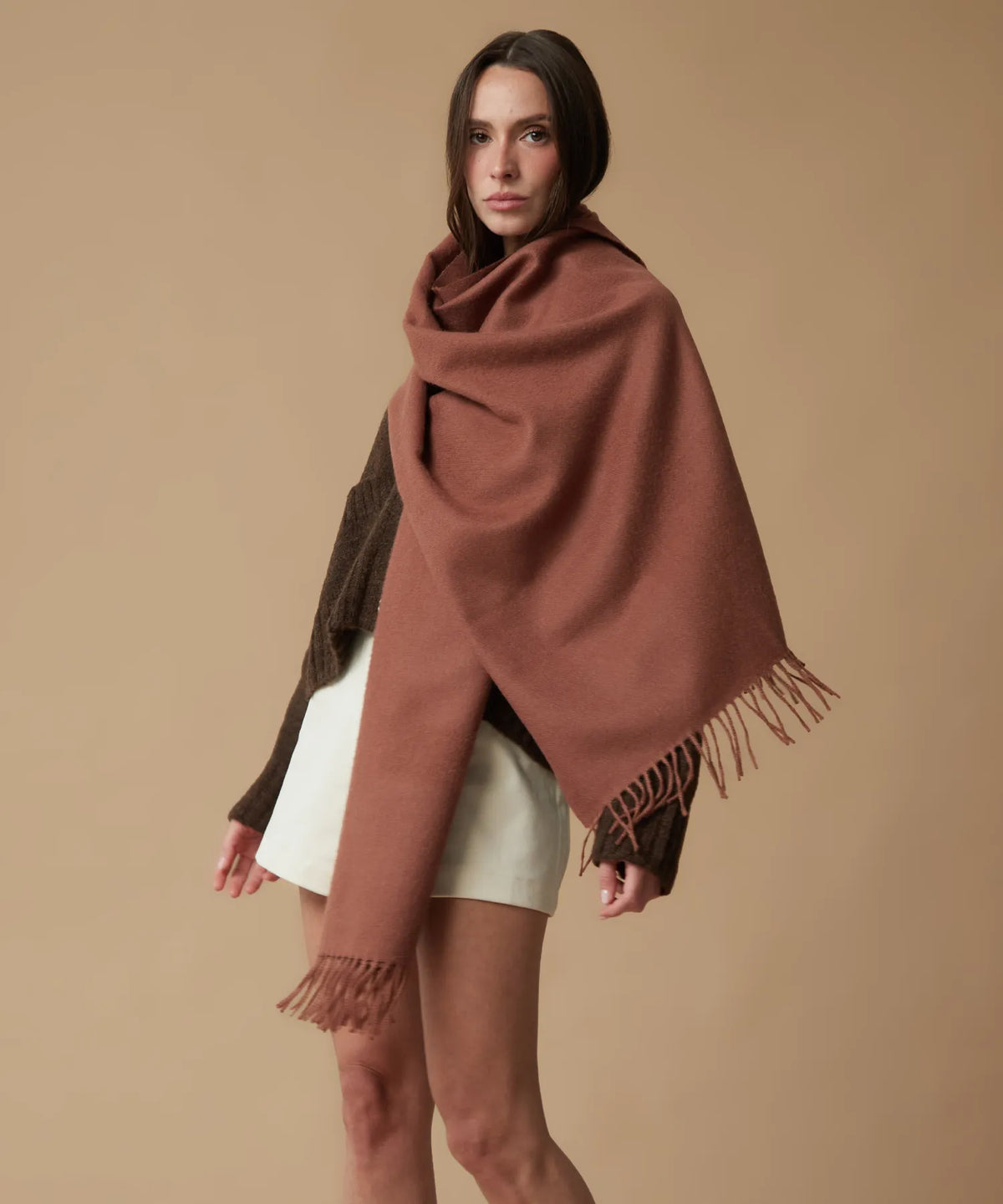 A woman modeling a large rust-brown fringed wrap or shawl draped over her shoulders, worn over a dark brown sweater and white shorts, against a solid beige studio background.