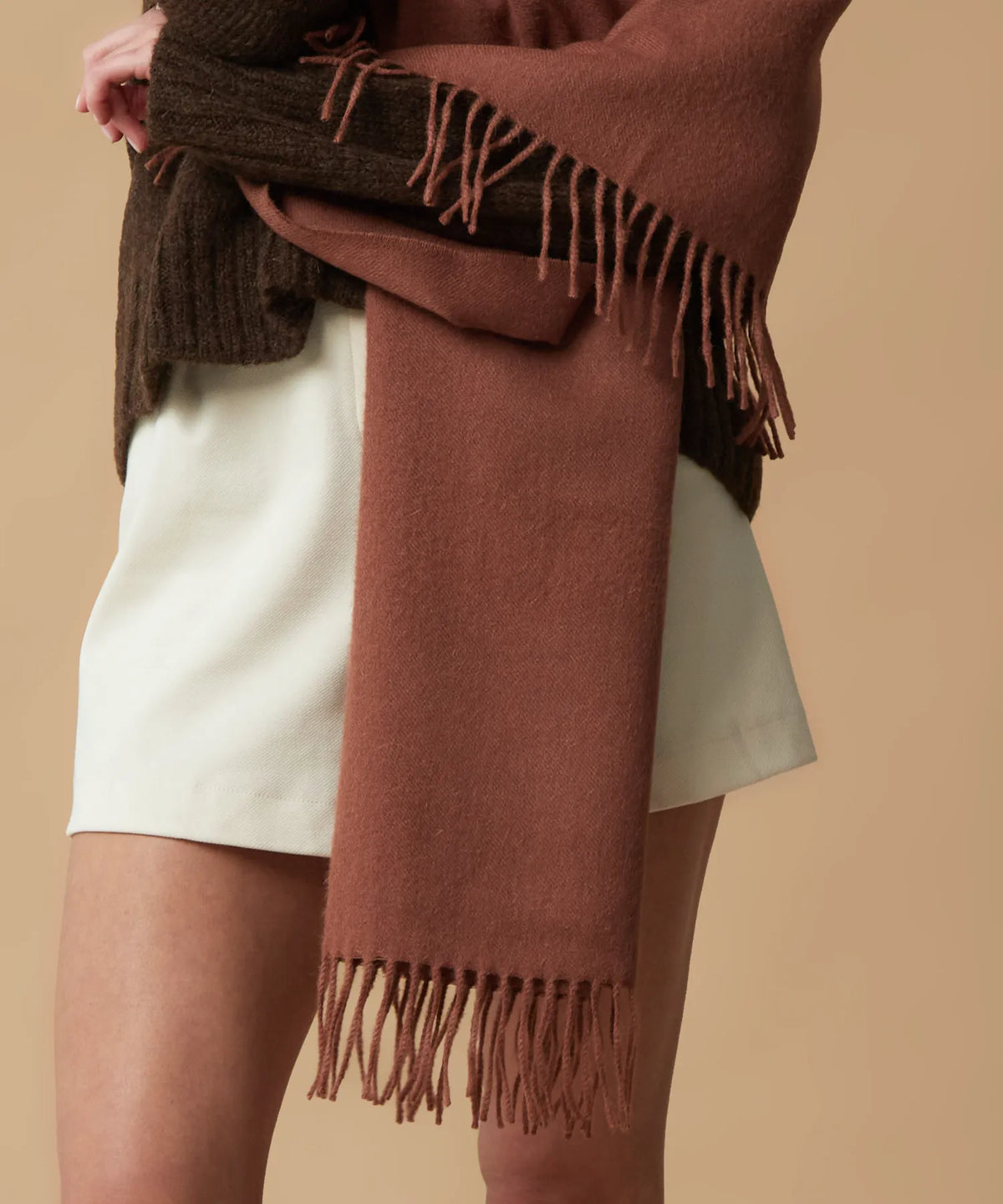 A close-up of a person wearing a rust-brown fringed wrap or shawl draped over a cream skirt and a dark brown knit top, against a solid beige background.