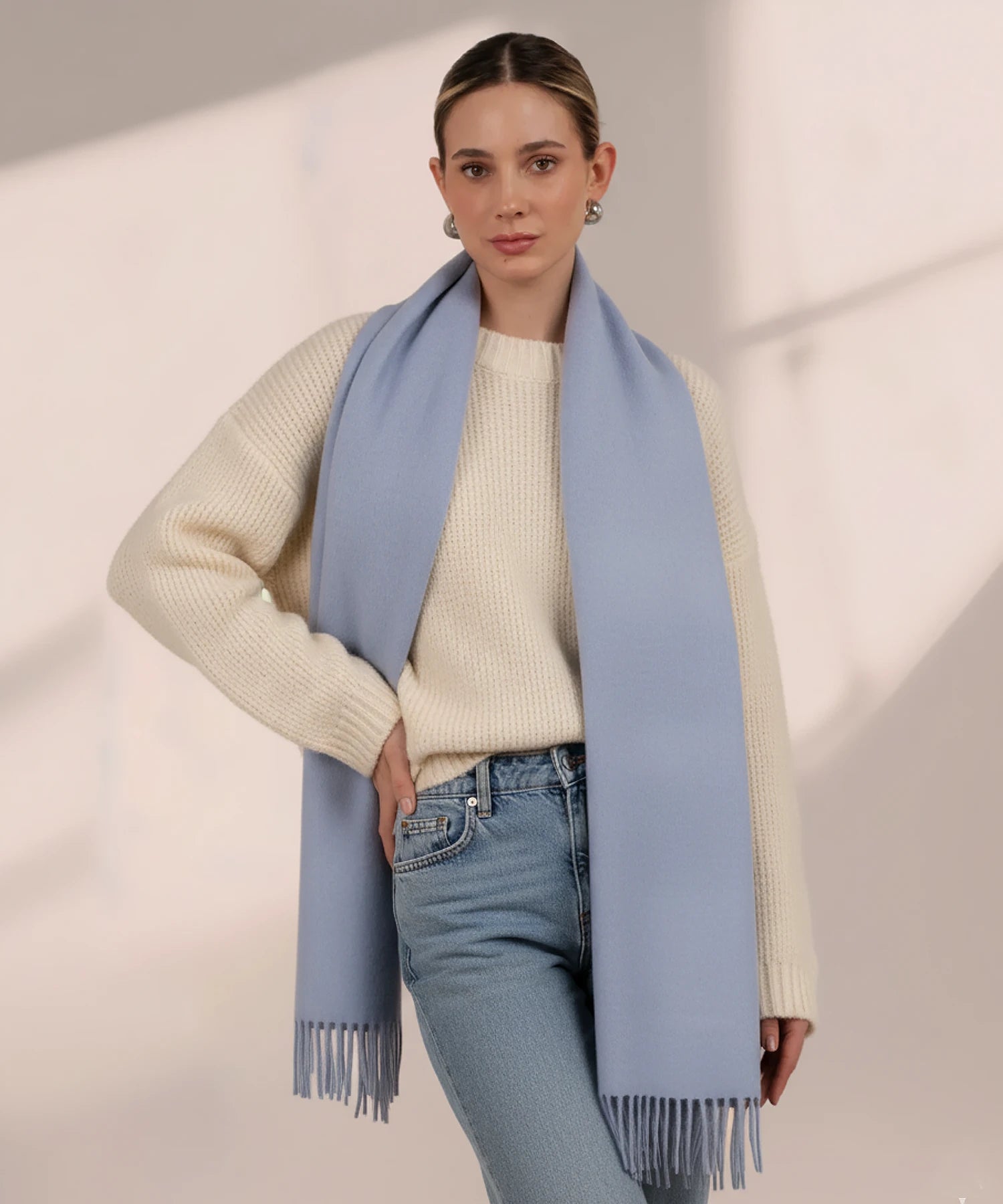 Woman wearing a light blue scarf, white sweater, and jeans on a plain background