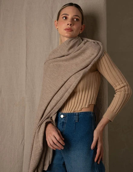 Woman wearing a beige sweater and blue jeans with a beige shawl draped over her shoulders against a plain background.