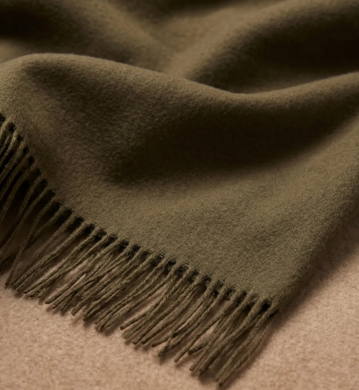 A close-up of a dark olive green scarf with fringed edges, draped over a light beige surface.