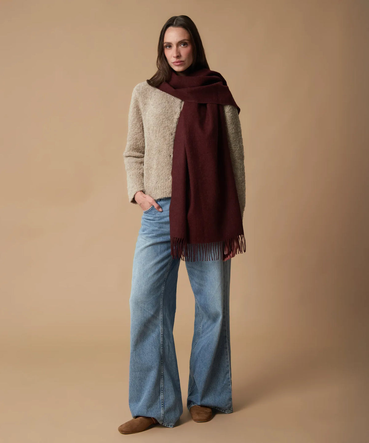 A woman stands against a plain beige studio background wearing a chunky knit color-block sweater in beige and olive green, light blue wide-leg jeans, and brown suede boots. She is wrapped in a large dark burgundy scarf with fringe.
