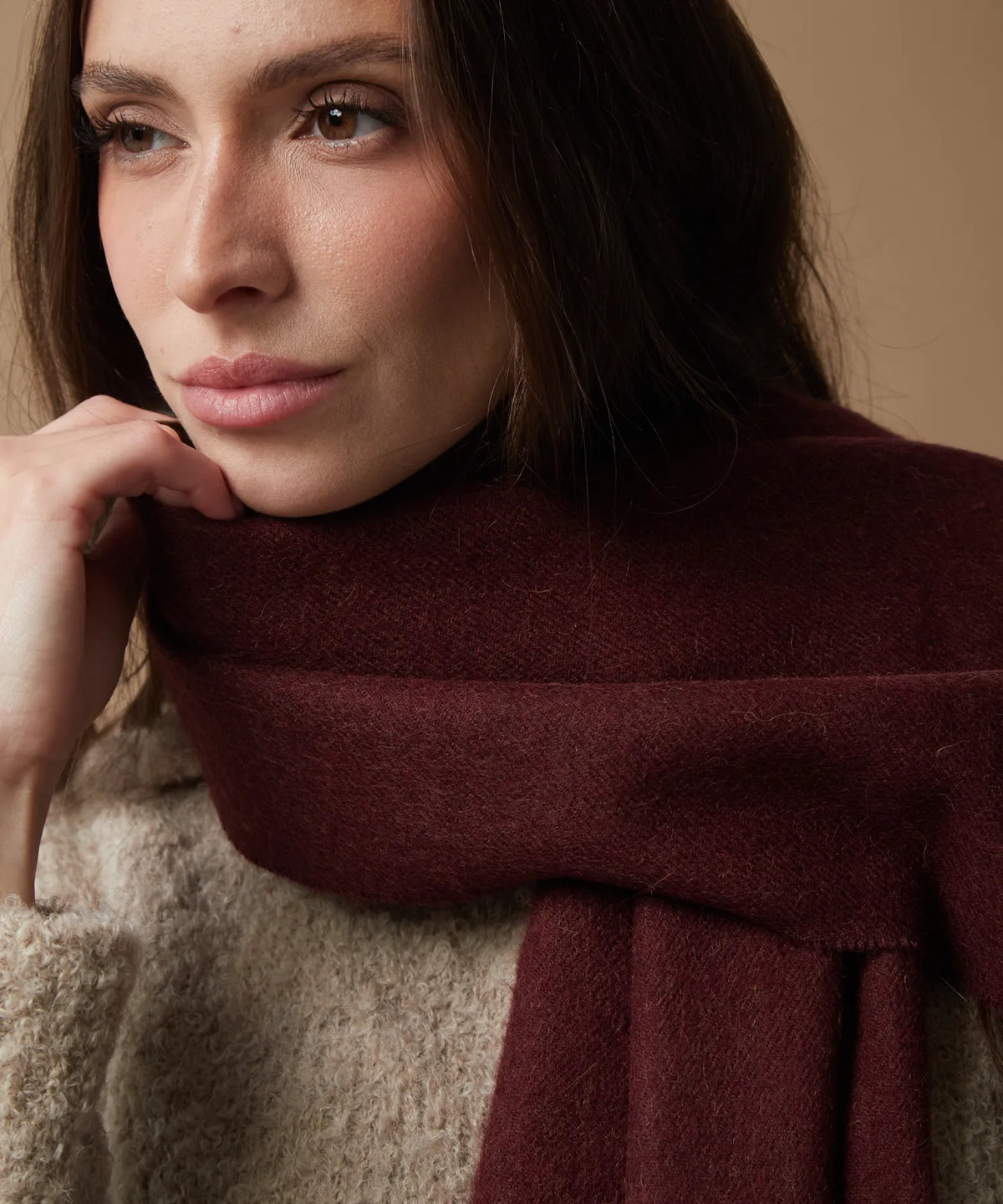 A close-up of a woman wearing a thick, textured burgundy alpaca scarf wrapped around her neck over a beige knit sweater, set against a warm beige background.