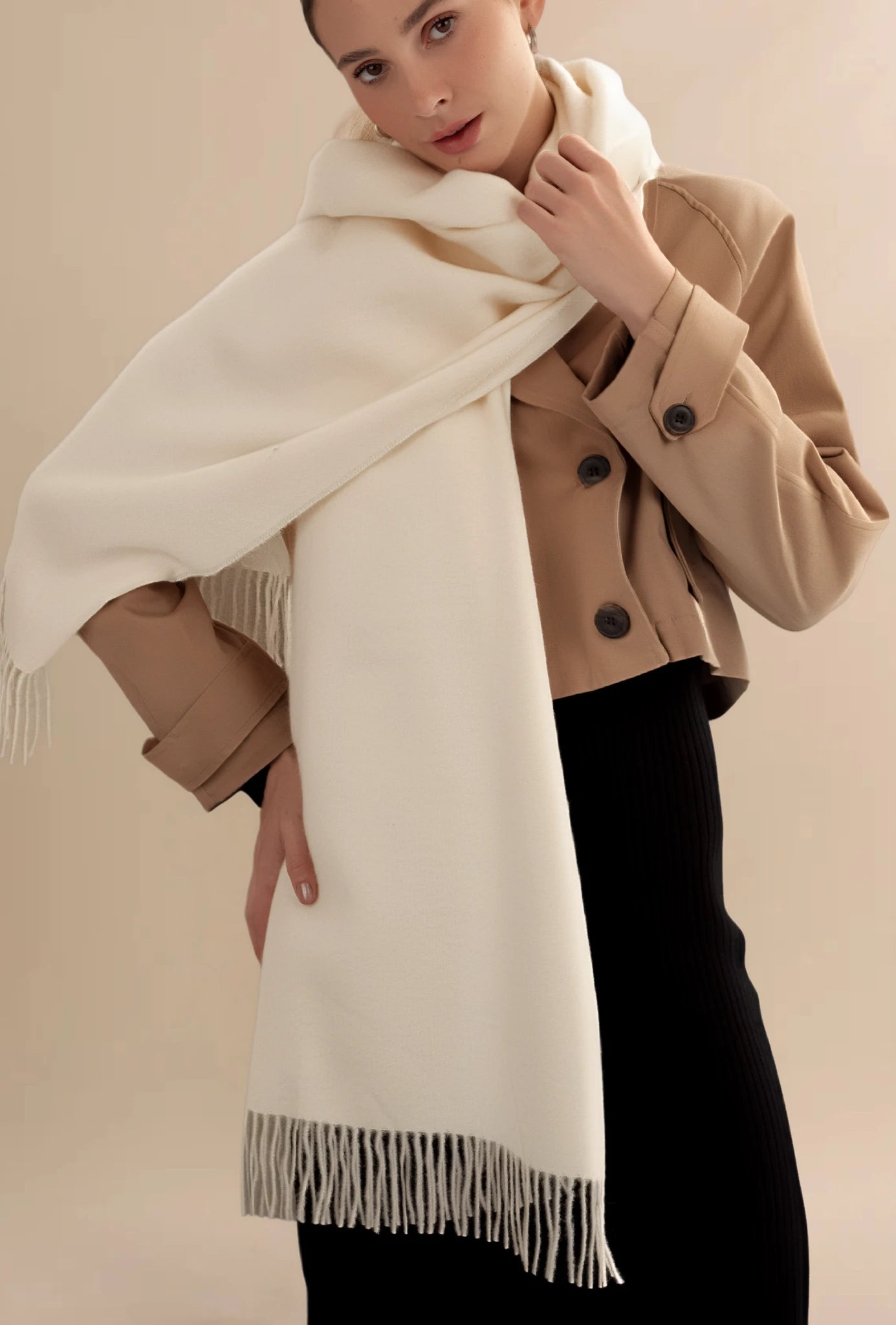 Woman wearing a beige coat and white scarf with fringe against a beige background