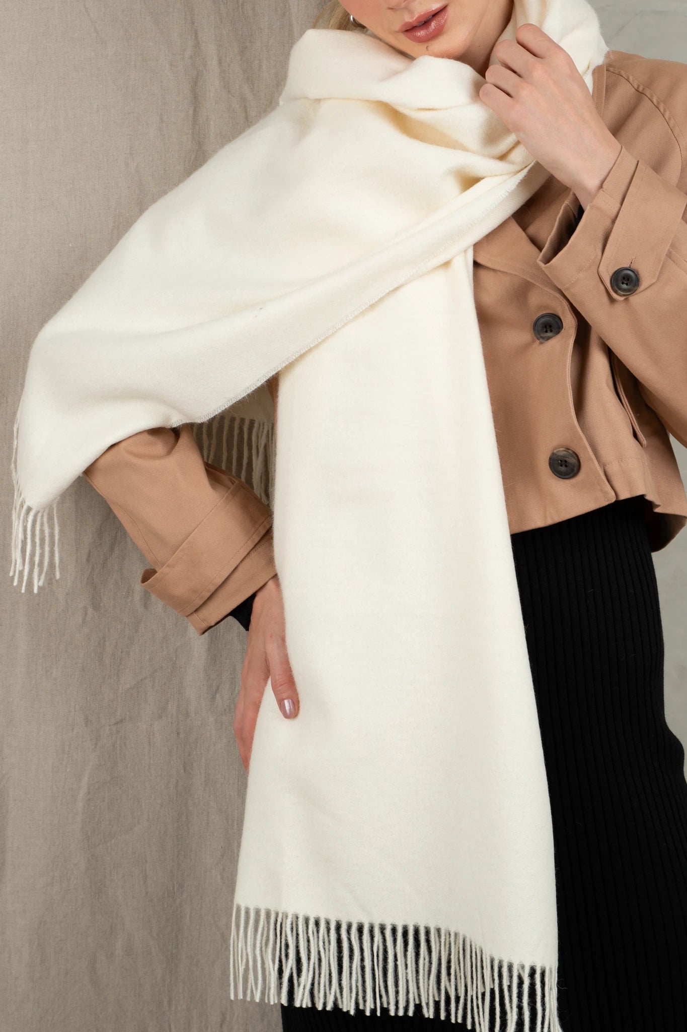 Person wearing a white scarf with a beige coat and black pants against a neutral background